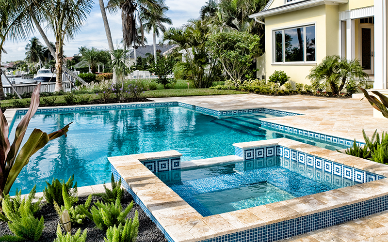 Rectangular swimming pool with a connected hot tub, surrounded by palm trees and greenery next to a yellow house with a tiled patio.