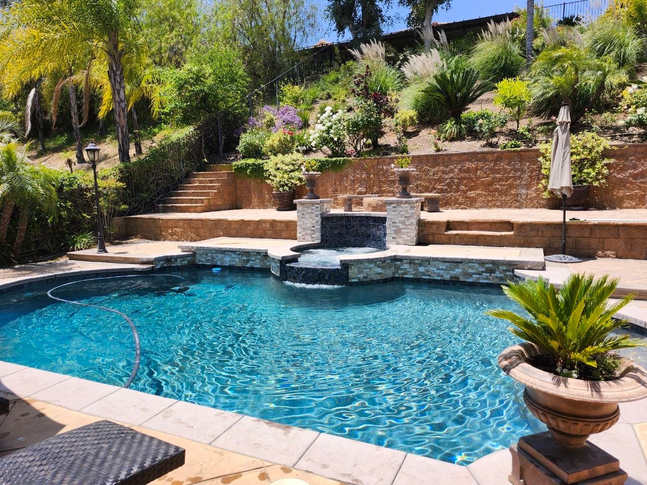 Backyard swimming pool with clear blue water, a built-in hot tub, stone waterfall feature, patio, and landscaped garden with various green plants and trees in the background.